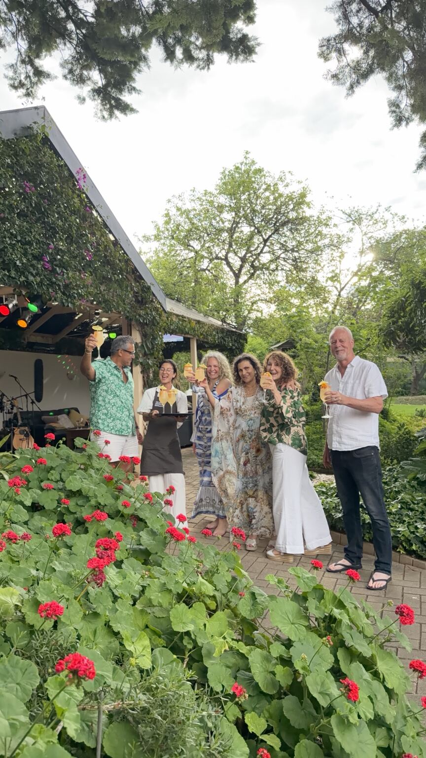 Friends raising golden cocktails on a garden path among red geraniums, with a server holding a tray beside them